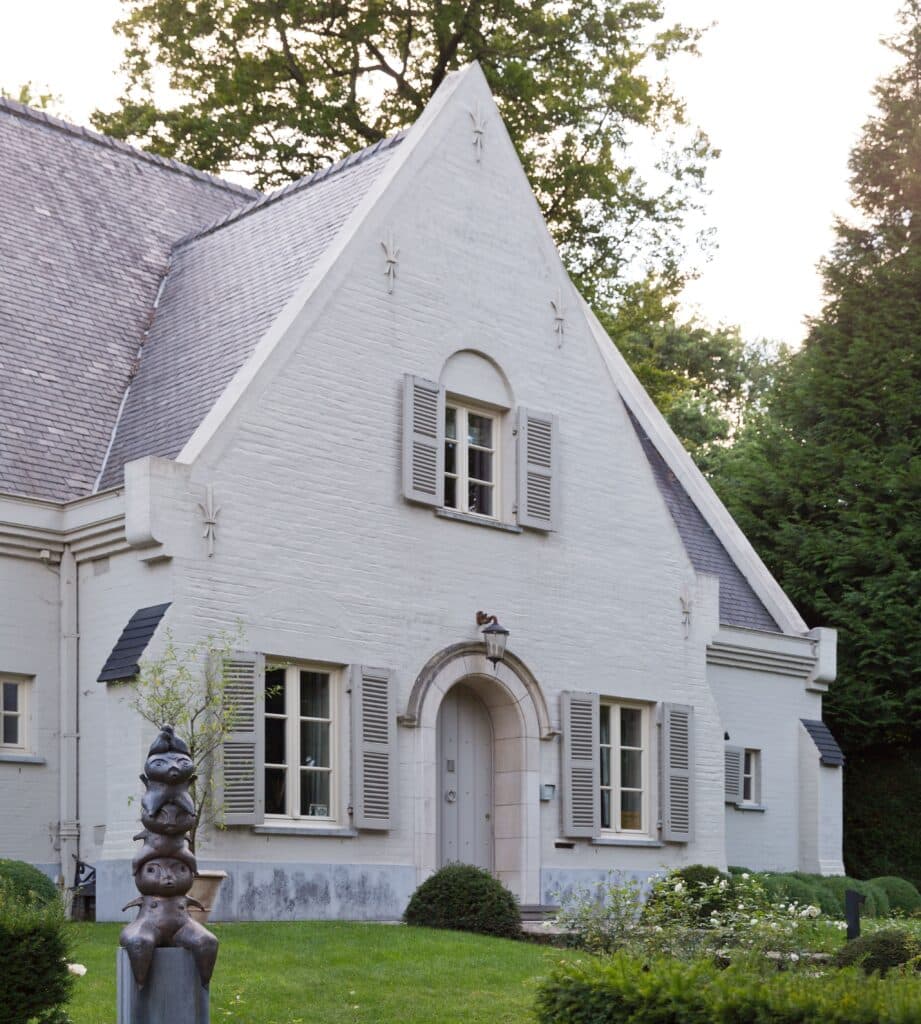 Maison traditionnelle avec une façade en briques blanches, équipée de volets en bois et de fenêtres en châssis bois Profel. Les éléments architecturaux, comme la porte en arc et les détails soignés, apportent du charme et du caractère à cette demeure. La statue dans le jardin et les arbustes bien entretenus ajoutent une touche artistique et naturelle à l'environnement.