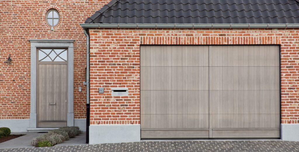 Entrée et garage d'une maison en briques rouges, avec une porte et une porte de garage en bois moderne, parfaitement assorties au style traditionnel de la façade. Les châssis bois Profel apportent une touche contemporaine tout en maintenant une harmonie visuelle avec les matériaux classiques. Les détails architecturaux, tels que la petite fenêtre ronde et la finition de la porte, ajoutent du caractère à cette propriété.