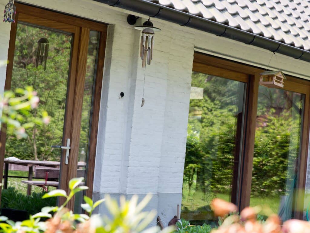 Façade d'une maison lumineuse avec de grandes baies vitrées en châssis bois Profel, s'ouvrant sur un espace verdoyant. Les cadres en bois ajoutent une touche naturelle et chaleureuse à l'architecture, tandis que les fenêtres offrent une belle connexion visuelle avec l'extérieur. Les éléments décoratifs, comme les carillons et la petite mangeoire suspendue, créent une ambiance paisible et conviviale.