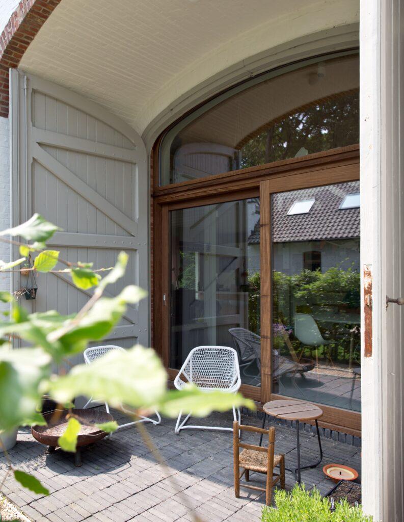 Terrasse abritée avec une grande baie vitrée en châssis bois Profel, s'ouvrant sur un espace extérieur convivial. La forme arrondie de la fenêtre apporte du caractère à cette façade traditionnelle, tandis que les châssis bois Profel offrent une isolation performante et une finition élégante. L’association du bois et des matériaux naturels crée une ambiance chaleureuse et accueillante.