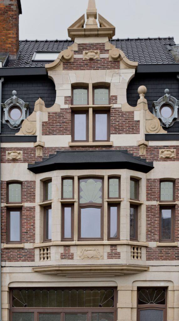 Façade richement décorée d'un bâtiment historique, avec des châssis bois Profel parfaitement intégrés à l'architecture ornementale en pierre et briques rouges. Les fenêtres en châssis bois, ornées de vitraux, respectent l'authenticité du style tout en offrant une performance énergétique moderne. Les détails sculpturaux et la baie vitrée en saillie confèrent à ce bâtiment un charme unique.