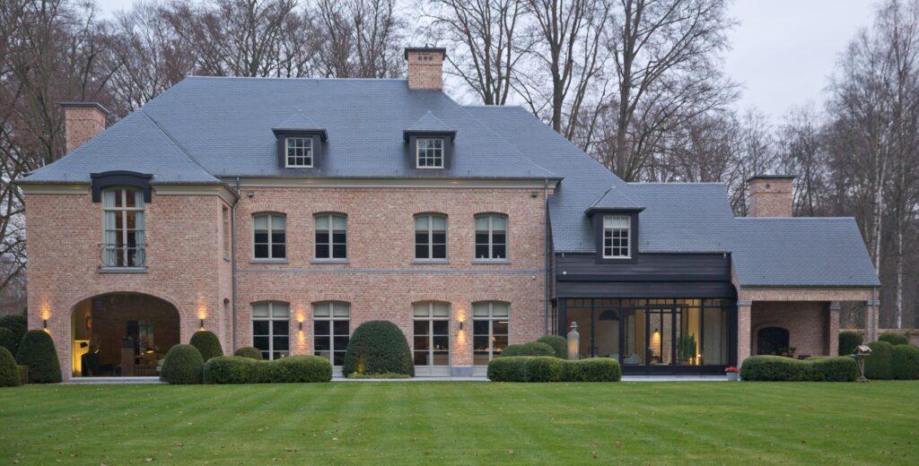 Grande maison de style traditionnel avec une façade en briques rouges et des châssis bois Profel, combinant élégance et performance énergétique. Les nombreuses fenêtres en châssis bois Profel apportent une luminosité naturelle à l'intérieur, tout en garantissant une isolation optimale. Le toit en ardoise et les éléments architecturaux raffinés soulignent l'authenticité et le caractère intemporel de la demeure.