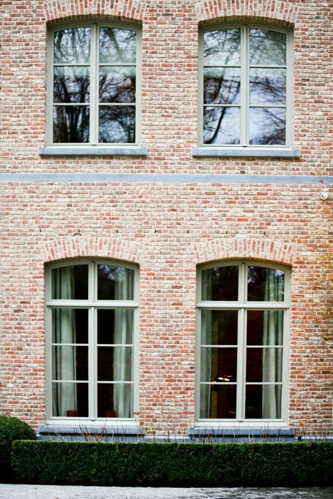 Façade en briques rouges ornée de grandes fenêtres équipées de châssis bois Profel, alliant esthétique et performances énergétiques. Les fenêtres cintrées au rez-de-chaussée et rectangulaires à l’étage apportent une élégance intemporelle à la structure. Les châssis bois Profel sont parfaitement intégrés à cette architecture traditionnelle, offrant durabilité et confort thermique