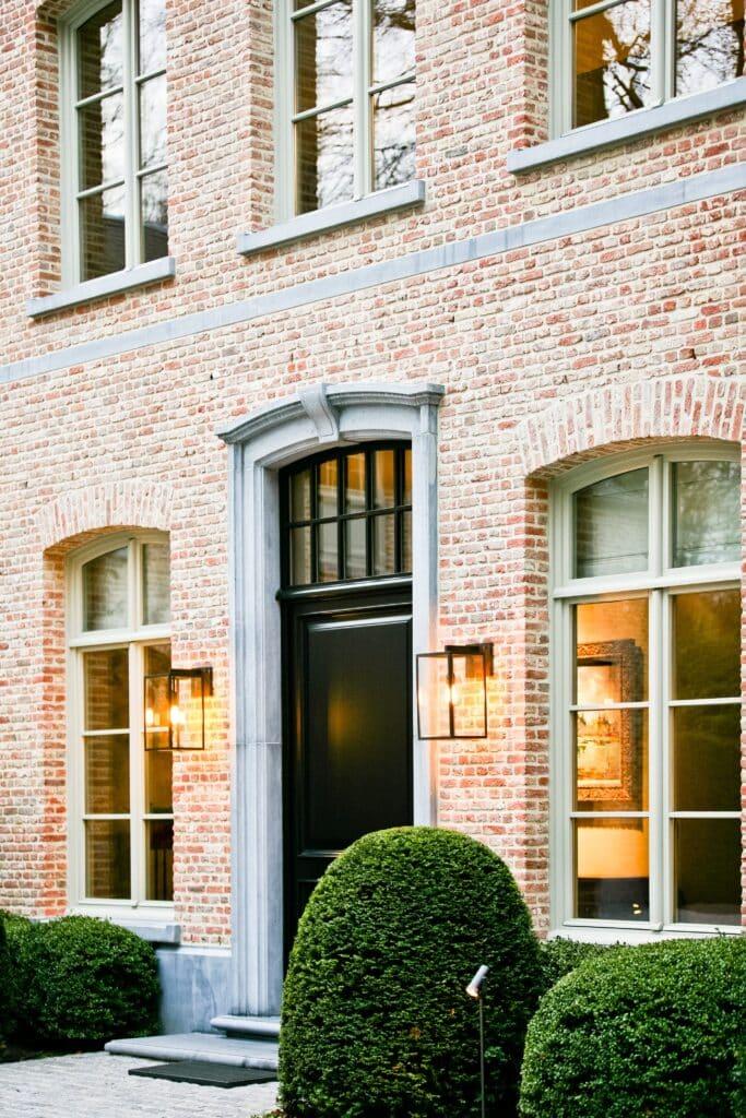 Entrée principale élégante d'une maison traditionnelle avec des châssis bois Profel, mettant en valeur l'authenticité et le charme de la façade en briques rouges. Les fenêtres en châssis bois Profel assurent un éclairage naturel optimal tout en offrant une isolation efficace. La porte d'entrée, encadrée par de superbes éléments en pierre et illuminée par des appliques modernes, ajoute une touche de sophistication à cette propriété.