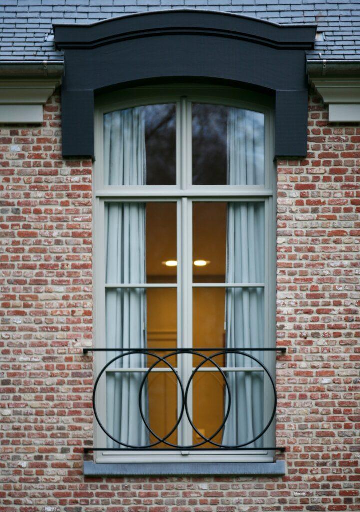 Fenêtre en châssis bois Profel dans une façade en briques rouges, avec une finition soignée et un garde-corps en fer forgé ornemental. La fenêtre cintrée ajoute une touche d'élégance à l'ensemble, tout en offrant une isolation optimale et une vue imprenable. Le châssis bois Profel s'intègre parfaitement au style traditionnel de la maison, combinant esthétique et durabilité.