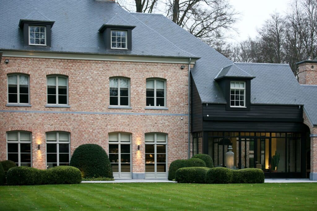 Façade d'une maison élégante avec des châssis en bois Profel, combinant harmonieusement des matériaux modernes et traditionnels. Les grandes fenêtres en châssis bois Profel offrent une luminosité naturelle optimale tout en garantissant une isolation thermique et acoustique exceptionnelle. Le style classique de la maison, avec ses briques rouges et ses toits mansardés, est magnifiquement mis en valeur par la qualité des châssis bois, soulignant le savoir-faire artisanal et la robustesse des matériaux. Les haies bien taillées et la pelouse verdoyante complètent l'aspect soigné et raffiné de cette propriété.