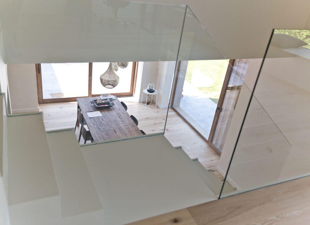 Vue d'intérieur moderne et lumineux avec un escalier en verre transparent donnant sur une salle à manger spacieuse. Les grandes baies vitrées équipées de châssis bois Profel offrent une belle ouverture sur l'extérieur, inondant l'espace de lumière naturelle. L'élégance du bois et la modernité du verre se marient harmonieusement pour créer une atmosphère à la fois chaleureuse et contemporaine