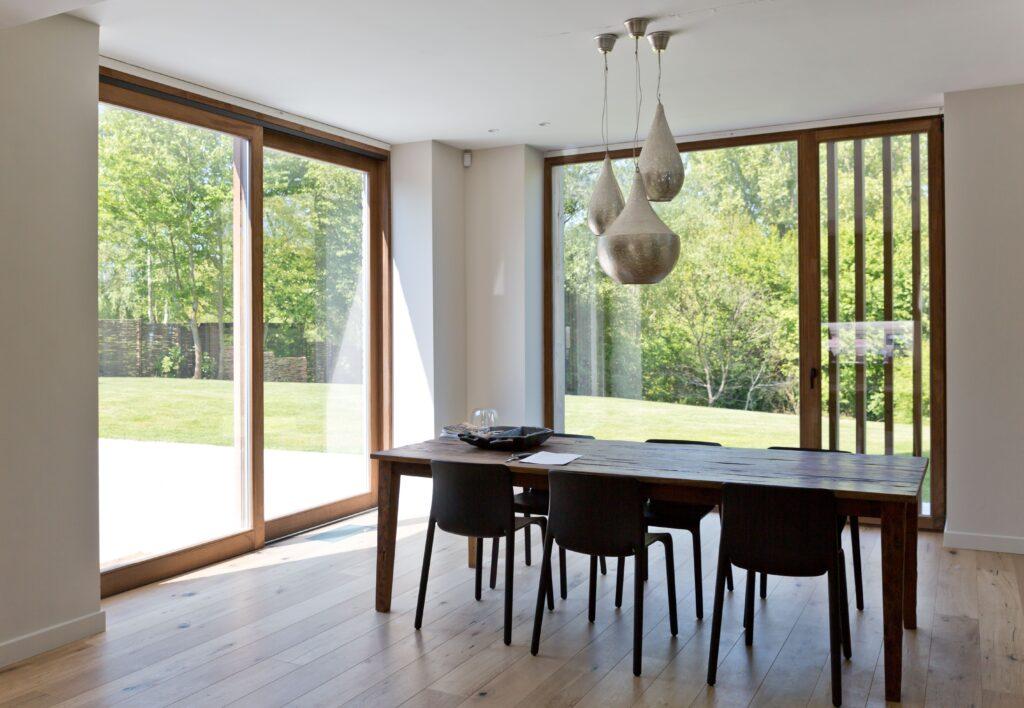 Salle à manger moderne et lumineuse, avec de grandes baies vitrées en châssis bois Profel offrant une vue dégagée sur le jardin. Les fenêtres en bois apportent une chaleur naturelle à l'espace tout en permettant une connexion fluide entre l'intérieur et l'extérieur. La lumière naturelle inonde la pièce, mettant en valeur le parquet en bois et le mobilier contemporain.