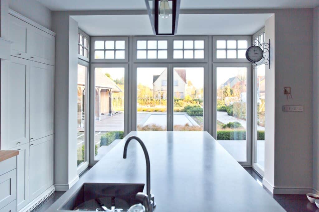 Cuisine moderne et lumineuse avec une large baie vitrée en châssis bois Profel, offrant une vue dégagée sur le jardin et inondant la pièce de lumière naturelle. Les fenêtres aux carreaux supérieurs ajoutent une touche classique à l'esthétique contemporaine de l'espace. L'îlot central avec évier et robinetterie en acier inoxydable crée un espace de travail fonctionnel et élégant.